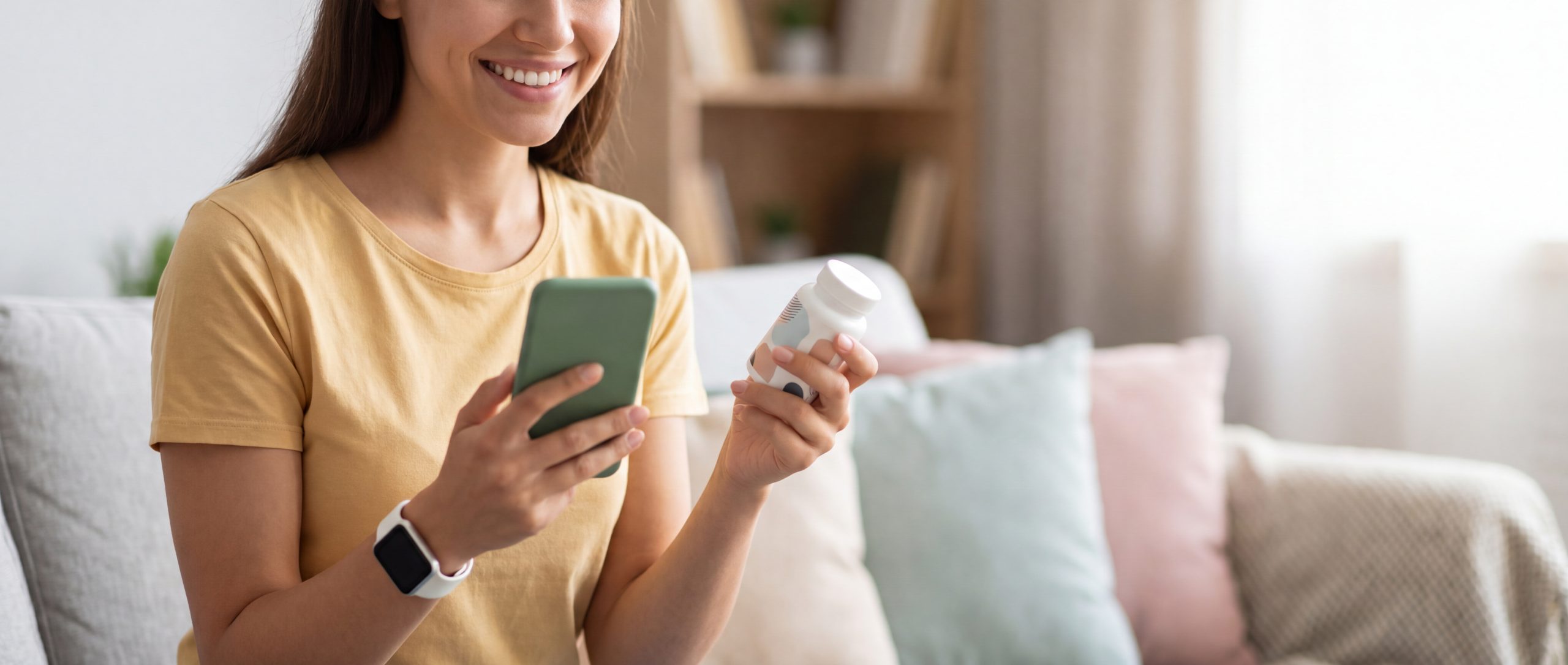Woman holding pill bottle in one hand and phone in the other sitting on a couch in the living room